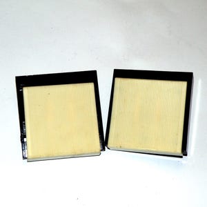 May include: Two square black frames with a light yellow, vertically-lined interior. The frames are side-by-side against a white background. The frames appear to be filters or components of a larger device.