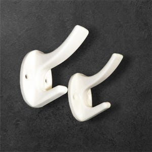 May include: Two white wall-mounted hooks with a smooth, glossy finish. Each hook features a curved upper arm and a smaller lower arm, with visible screw holes on the mounting plate for attachment to a flat surface.