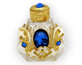 May include: A small, clear glass bottle with a gold-colored top and intricate gold and white detailing. The bottle has a large blue gemstone in the center and smaller blue gemstones around the edges.