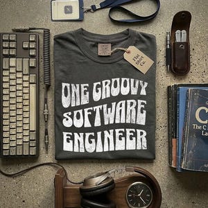 Könnte beinhalten: Draufsicht mit einem dunkelgrauen T-Shirt mit dem weißen Text "ONE GROOVY SOFTWARE ENGINEER". Das Bild enthält eine Tastatur, Kopfhörer, eine Uhr, Bücher und ein Multitool.