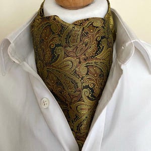 Puede incluir: Un corbata ascot estampada en tonos dorados, marrones y negros, con un diseño de cachemira. La corbata se lleva sobre una camisa blanca con cuello, con el botón superior abrochado. La tela tiene un ligero brillo.