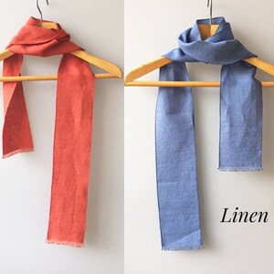 May include: Two linen scarves hanging on a wooden hanger. The scarf on the left is a bright orange color. The scarf on the right is a light blue color. The text "Linen" is printed below the scarves.