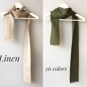 May include: Two linen scarves hanging on white hangers. The scarf on the left is a light beige color. The scarf on the right is a dark green color. The text "Linen" is below the beige scarf and the text "26 colors" is below the green scarf.