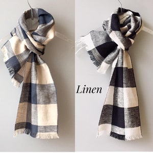 May include: Two linen scarves, one in navy and beige, the other in black and beige, are displayed on hangers. Both scarves have a checkered pattern and fringed edges. The word "Linen" is printed on the image.