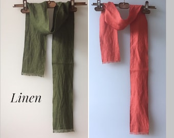 Könnte beinhalten: Zwei Leinenschals hängen an einem Holzbügel. Der Schal links ist dunkelgrün und der Schal rechts ist korallenfarben. Beide Schals haben einen Fransenrand.