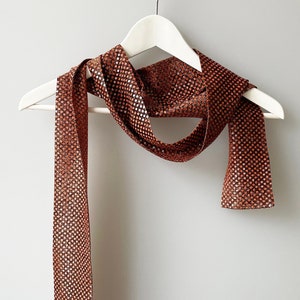 May include: A brown sequined scarf with a geometric pattern hangs on a white hanger against a white background.
