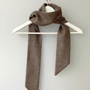 May include: A brown knitted scarf hanging on a white hanger. The scarf is long and narrow with a pointed end.