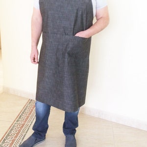 Puede incluir: Un delantal de mezclilla gris con un bolsillo grande en la parte delantera. El delantal tiene correas ajustables y se usa sobre una camiseta blanca.