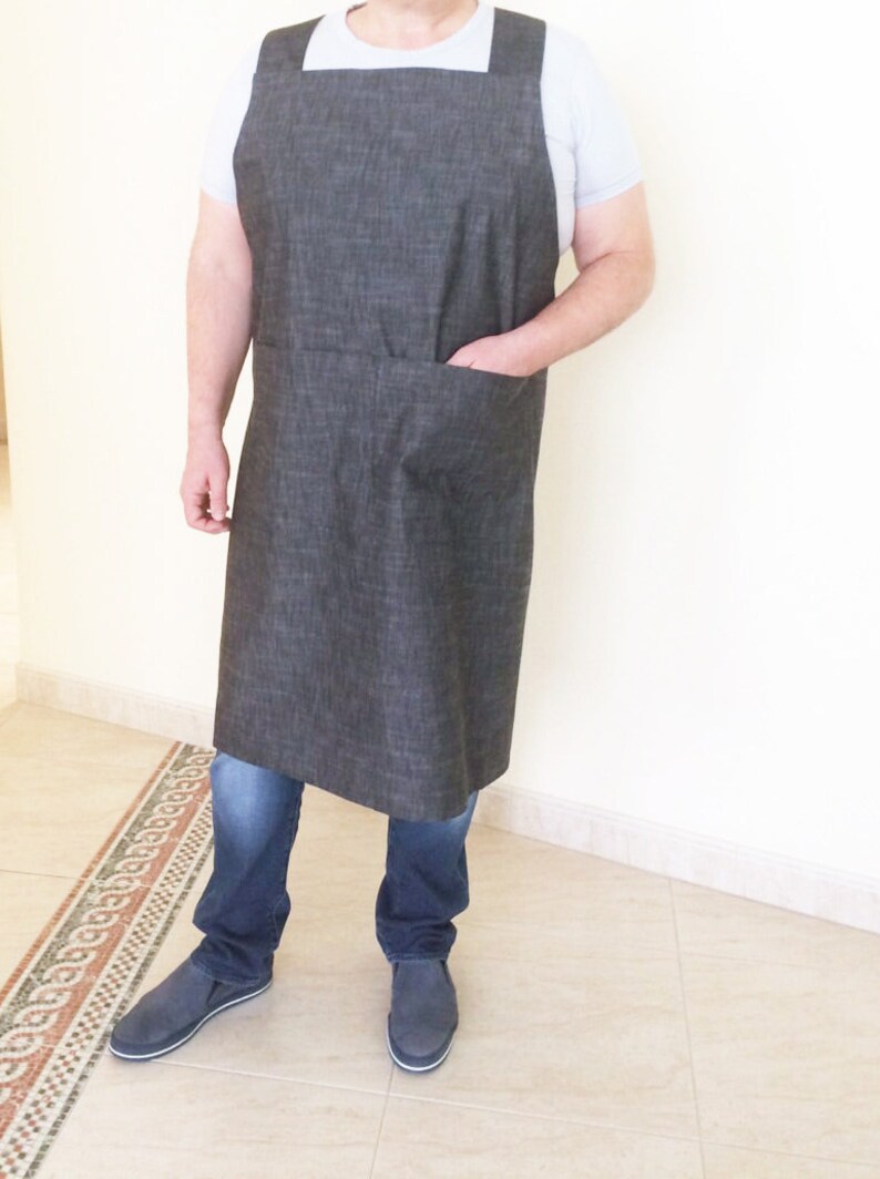 Puede incluir: Un delantal de mezclilla gris con un bolsillo grande en la parte delantera. El delantal tiene correas ajustables y se usa sobre una camiseta blanca.