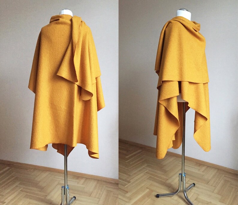 May include: A mustard yellow wool cape draped over a mannequin. The cape is asymmetrical and has a flowing design.