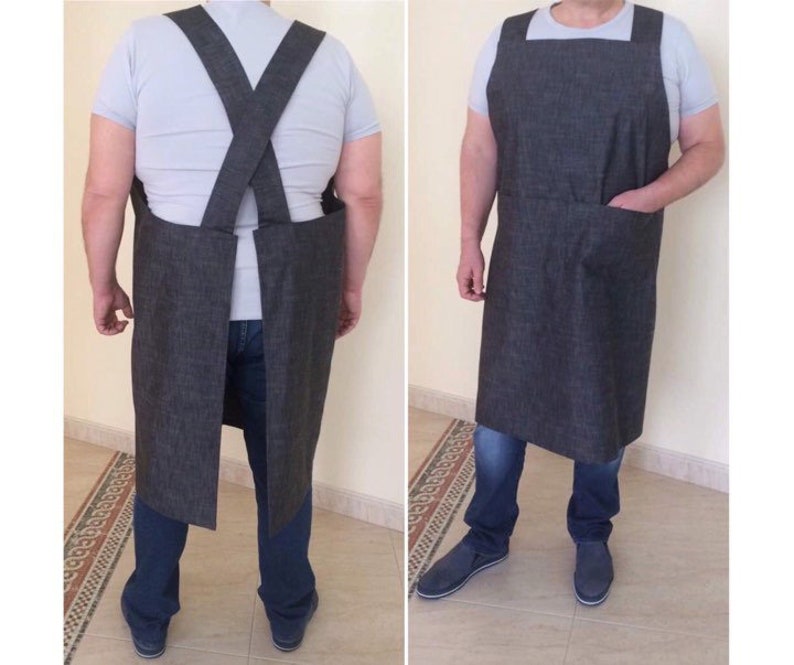 Puede incluir: Un delantal de mezclilla gris oscuro con tirantes ajustables y un bolsillo grande. El delantal se usa sobre una camiseta blanca y jeans azules.
