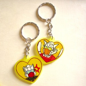 May include: Two yellow heart-shaped keyrings with cartoon robot characters. The keyring on the left features a robot with a red face and a yellow star. The keyring on the right features a robot with an orange face and a white and red body.