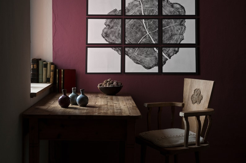 May include: A wooden table with a dark brown finish sits in a room with a burgundy wall. Three small ceramic vases are on the table, along with a bowl of dried pinecones. A wooden chair with a carved back is in the foreground. A framed print of a tree trunk cross-section hangs on the wall.