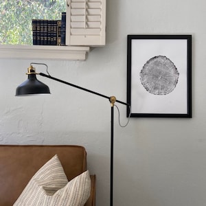 Puede incluir: Una impresión en blanco y negro de una sección transversal de tronco de árbol en un marco negro cuelga de una pared blanca. Una lámpara de pie negra con un acento dorado está en primer plano. Un sofá de cuero marrón con un cojín estampado en blanco y negro está en la esquina inferior izquierda.