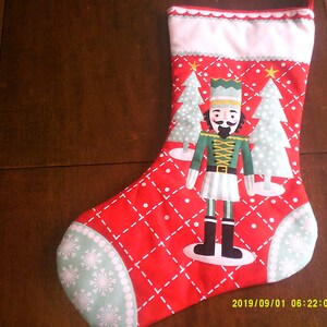 Op de afbeelding: Rode kerstsok met een witte boord en een groen-witte notenkrakerontwerp. De sok heeft een notenkraker die een staf vasthoudt, staande tussen twee groen-witte kerstbomen. De sok heeft een wit-groen sneeuwvlokkenpatroon aan de teen.