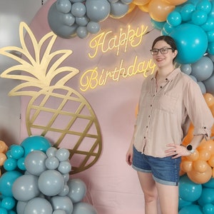 Può includere: Una donna si trova davanti a uno sfondo rosa con un ritaglio di ananas dorato e un cartello al neon che dice "Buon compleanno". Lo sfondo è decorato con un arco di palloncini blu, grigi e arancioni.
