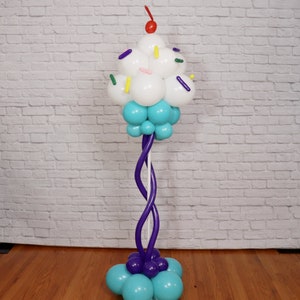 Pu&ograve; includere: Una scultura di palloncini a forma di cupcake con glassa bianca, granella blu e una ciliegia rossa in cima. Il cupcake &egrave; attaccato a una colonna di palloncini viola.