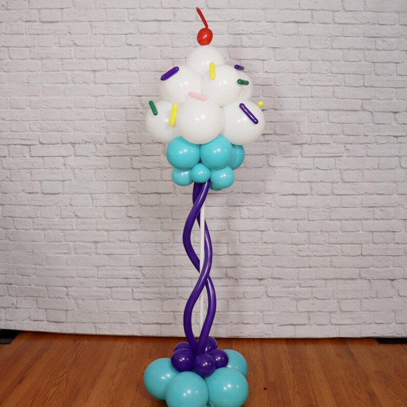 Pu&ograve; includere: Una scultura di palloncini a forma di cupcake con glassa bianca, granella blu e una ciliegia rossa in cima. Il cupcake &egrave; attaccato a una colonna di palloncini viola.