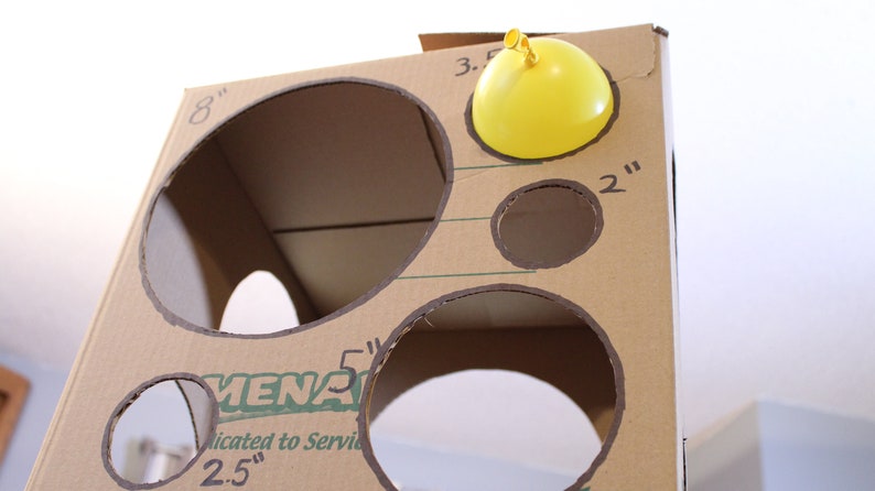 May include: A cardboard box with various sized holes cut out. The holes are labelled with measurements in inches. A yellow balloon is taped to the top of the box.