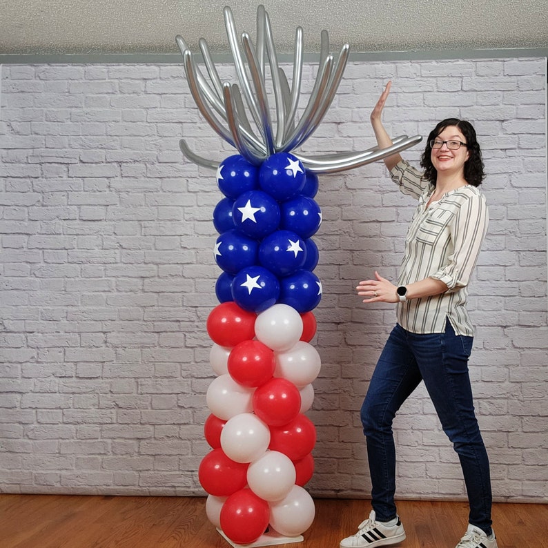 May include: A red, white and blue balloon column with a silver balloon arch on top. The column is made of red, white and blue balloons arranged in a pattern to resemble the American flag. The arch is made of silver balloons arranged in a fan shape. The column is standing on a white base.