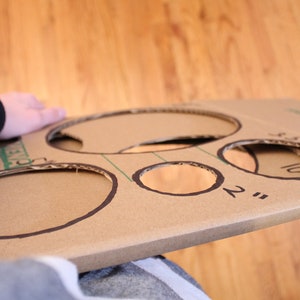 May include: A cardboard game board with five circles cut out. The circles are labelled with different sizes in inches: 2", 3", 4", 5", and 10".