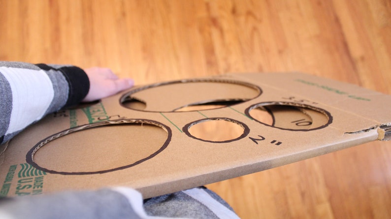 May include: A cardboard game board with five circles cut out. The circles are labelled with different sizes in inches: 2", 3", 4", 5", and 10".