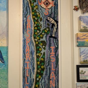 Peut inclure: Une œuvre d'art en point d'aiguille encadrée représentant une carte de l'île de Carrillia. L'île est délimitée en vert et entourée d'eau bleue. Le mot "ISLAND" est écrit verticalement sur le côté droit de la carte en lettres roses. Le mot "CARRILLIA" est écrit verticalement sur le côté gauche de la carte en lettres roses. Une rose des vents se trouve dans le coin supérieur gauche de la carte.