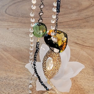May include: A multi-strand necklace with black, white and gold beads. The necklace features a large, faceted, green and gold gemstone pendant.