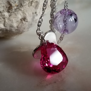 May include: A silver necklace with a large, heart-shaped, faceted pink gemstone pendant. The necklace also features a small silver charm and a lavender bead. The chain is a delicate silver cable.