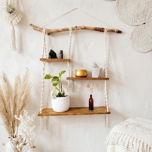 Peut inclure: Étagère suspendue en bois rustique à trois niveaux, suspendue par une corde blanche à une branche naturelle. Les étagères présentent des plantes en pot, une bougie et des bouteilles décoratives. Décoration bohème et naturelle.