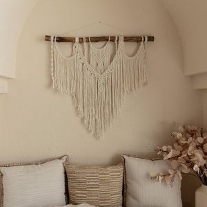 Peut inclure: Une suspension murale en macramé blanche avec une tringle en bois, suspendue au-dessus d'un canapé beige avec plusieurs coussins. Le canapé est dans une pièce aux murs blancs et une plante décorative.
