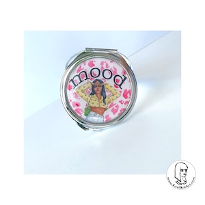 May include: A round, silver compact mirror with a pink leopard print background and the word "mood" in pink. The mirror features an illustration of a person wearing a yellow top and white jeans. A green purse is visible.