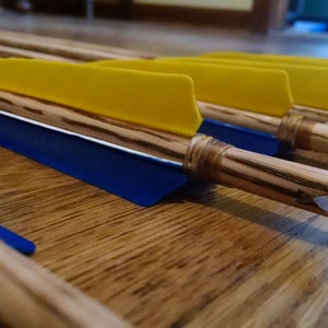 May include: Close-up of several wooden arrows with yellow and blue fletching. The arrows are lying on a wooden surface.