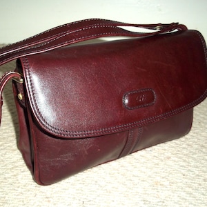 May include: A burgundy leather crossbody bag with a flap closure. The bag has a long adjustable strap and a small embossed logo on the front. The stitching is visible, and the hardware is gold-toned.