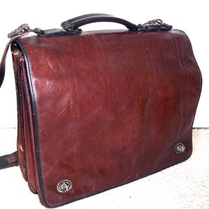 May include: A brown leather briefcase with a black handle and trim. The bag has a flap closure and a shoulder strap. The briefcase has two silver decorative accents on the front.