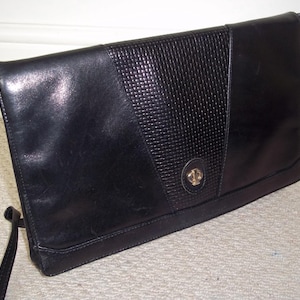 May include: Black leather shoulder bag with a textured flap and a gold-toned clasp. The bag has a long, thin strap and a rectangular shape. The textured section is a woven pattern. The bag is sitting on a light-colored carpet.