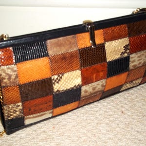 Op de afbeelding: Een rechthoekige patchwork clutch met een gouden kettingriem. De clutch heeft een verscheidenheid aan bruine, oranje en zwarte leren vierkanten. De boven- en onderranden zijn zwart en de sluiting is goudkleurig.
