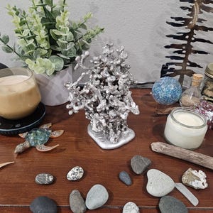 May include: A silver-colored metal sculpture with a textured, organic form sits on a wooden surface. Various decorative items surround it, including a small glass turtle, a candle, and several smooth stones. A small plant in a white pot is also visible.