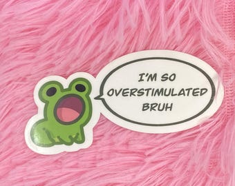 May include: A sticker featuring a cartoon frog with a wide open mouth, next to a speech bubble. The speech bubble has a black outline and contains the text "I'M SO OVERSTIMULATED BRUH". The background is pink.