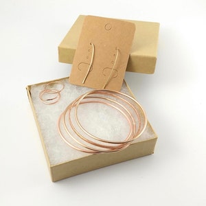 May include: A set of rose gold jewelry in a brown box. The set includes a pair of hoop earrings, a pair of drop earrings, and two rings.