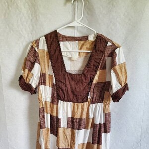 Puede incluir: Top de manga corta con diseño de patchwork en tonos marrón, beige y blanco. El top presenta un escote cuadrado con un ribete texturizado y cintura fruncida. La prenda está colgada de una percha blanca sobre un fondo blanco.