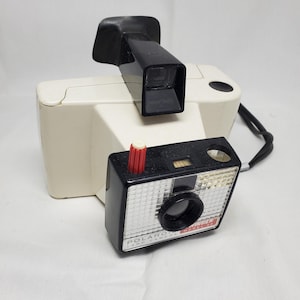 May include: A white Polaroid Land Camera Model 100 with a black lens and a red button. The camera has a black strap attached.