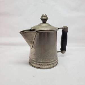 May include: A silver metal coffee pot with a black wooden handle and a decorative knob on the lid.