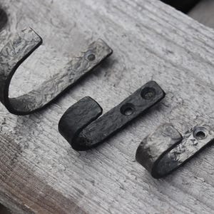 May include: Three handcrafted, dark gray metal hooks with a textured finish are arranged on a weathered wooden surface. The hooks vary in size and shape, with a curved design for hanging items.