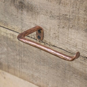 May include: A hammered copper toilet paper holder mounted on a wooden wall. The holder is shaped like a triangle with a rounded edge.