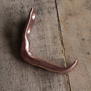 May include: A copper-colored metal hook with a curved design. The hook is mounted on a wooden surface.