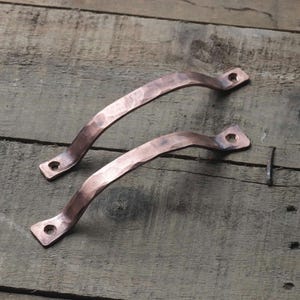 May include: Two copper-colored, curved cabinet handles with a hammered texture. Each handle has a rectangular base with two holes for mounting. The handles are displayed on a weathered wooden surface.