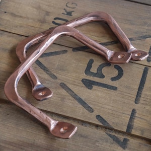 May include: Two copper-colored metal handles with a curved design. The handles are placed on a wooden surface with black stenciled numbers and text. The handles have a rustic, industrial aesthetic.
