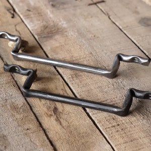 May include: Two metal cabinet pulls with a unique, wavy design. One is a dark, matte black, and the other is a lighter, silver-toned metal. The pulls are set against a rustic, wooden background, showcasing their handcrafted appearance.