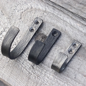 May include: Three dark gray metal hooks with a textured finish are arranged on a weathered wooden surface. Each hook has a rectangular base with pre-drilled holes for mounting and a curved hook. The hooks vary slightly in size.
